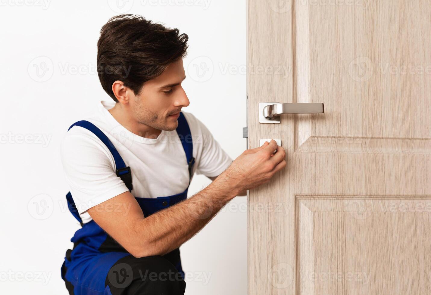 Repair Service Concept. Portrait of smiling handsome young locksmith in blue uniform installing door knob on front wooden entrance door at home or office workplace, isolated on white studio background photo