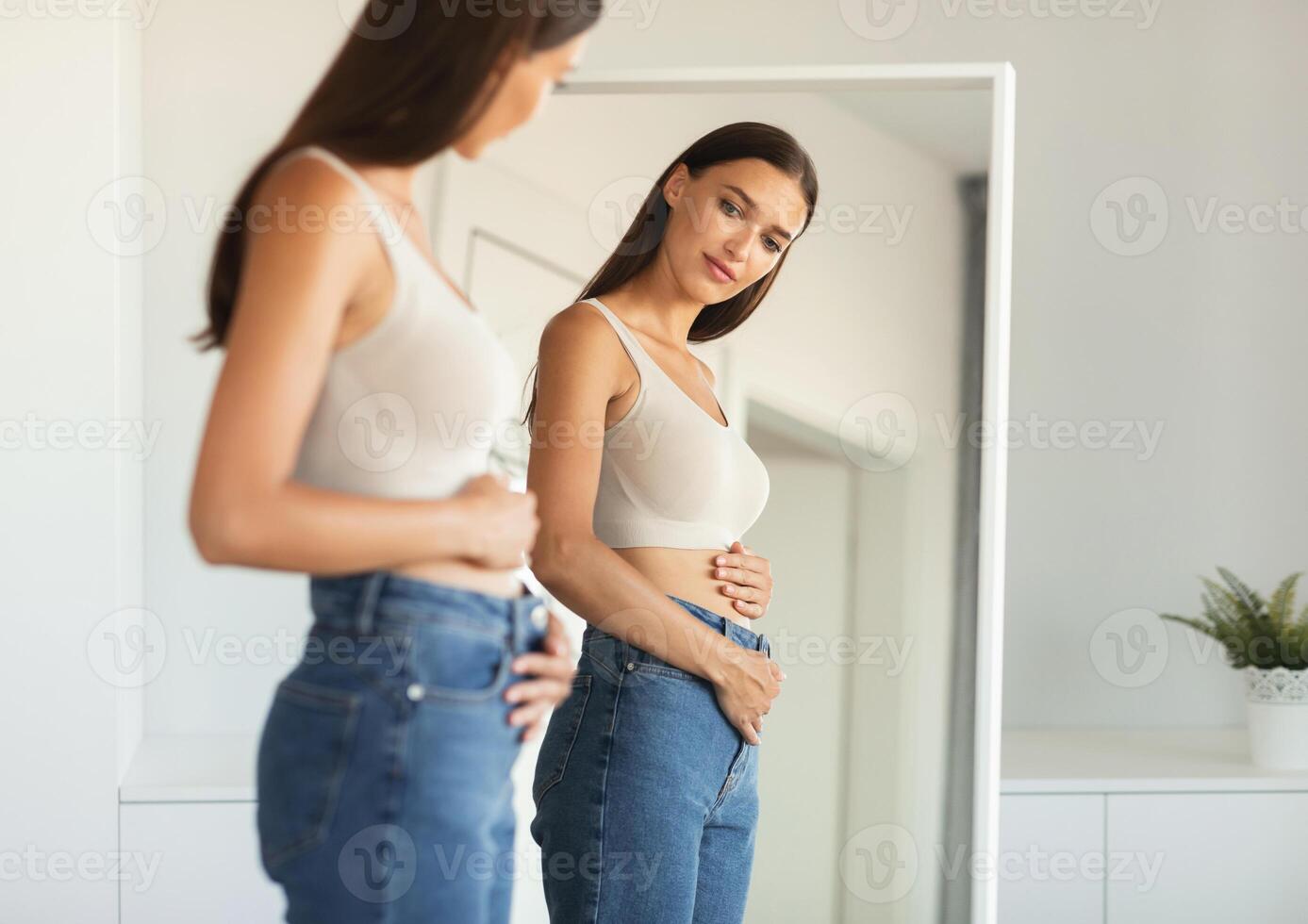 Young Woman Touching Flat Belly Expecting Baby And Enjoying First Months Of Pregnancy Posing Looking At Her Reflection In Mirror Wearing Skinny Jeans Standing At Home. Selective Focus photo