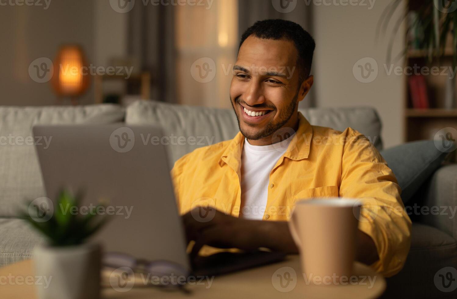 Smiling young black man working or communicating online, sitting on couch with laptop at home. Cheerful millennial guy freelancing, chatting on internet. Modern technologies in everyday life concept photo