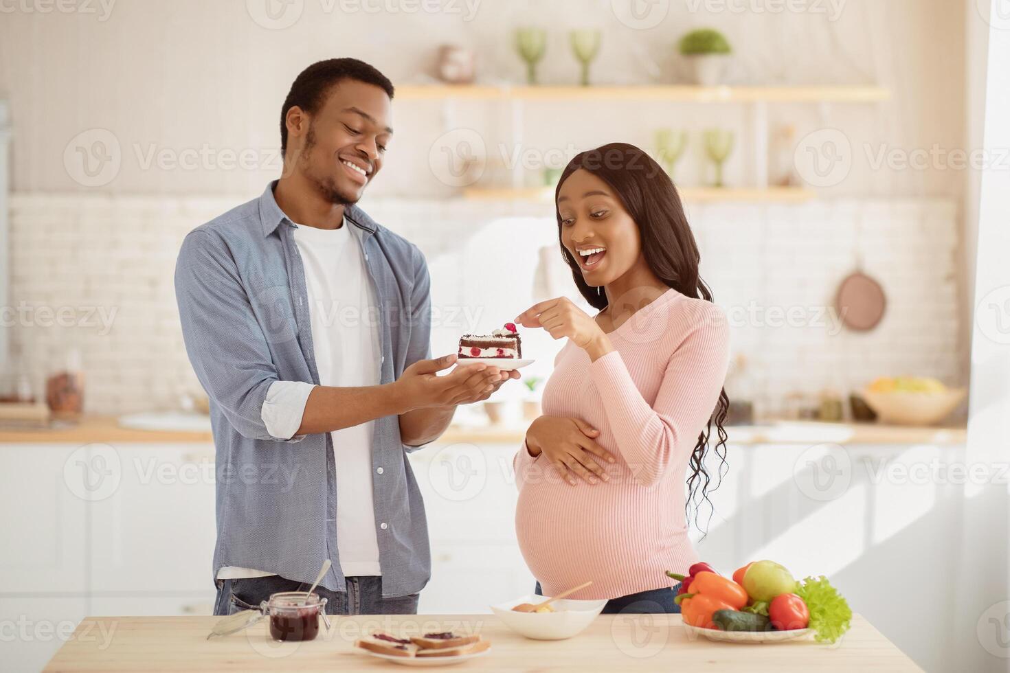 Handsome black man tempting his pregnant wife with piece of tasty cake in kitchen. African American husband offering dessert to his expectant girlfriend at home. Unhealthy nutrition during pregnancy photo