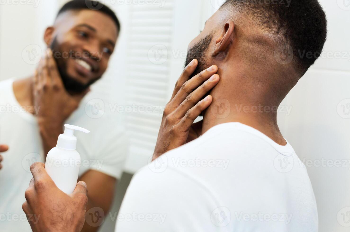 Facial care. Young black man applying moisturizer on beard in bathroom, looking in mirror photo