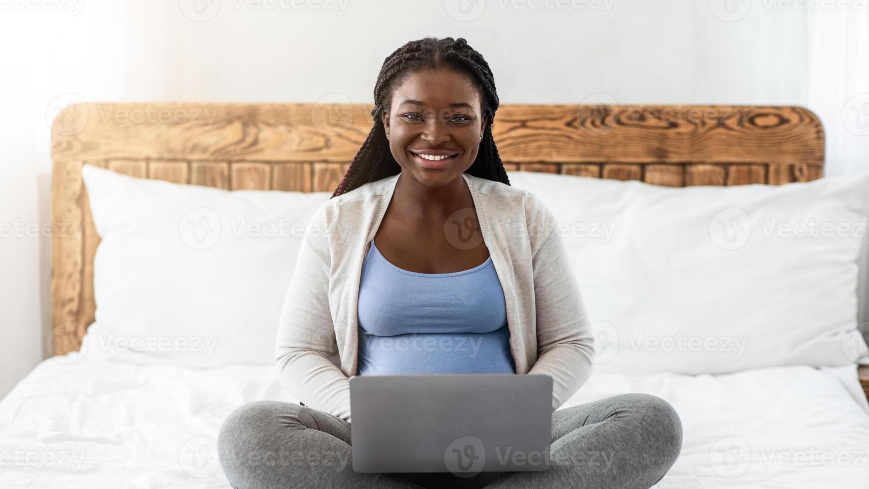 Remote Work For Pregnant Women. Happy Black Expectant Mother Using Laptop At Home, African American Lady Working Remotely While Sitting On Bed With Computer, Smiling At Camera, Copy Space photo