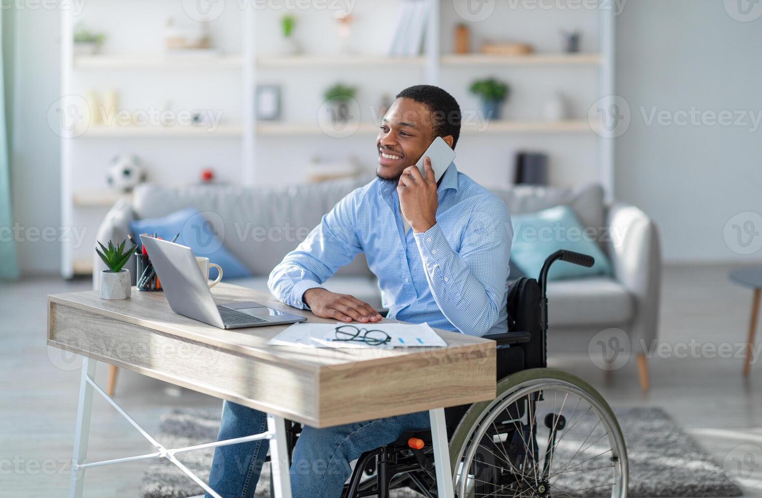 Joyful disabled black guy in wheelchair having phone conversation while working online on laptop from home. Young handicapped businessman talking with client on mobile phone photo