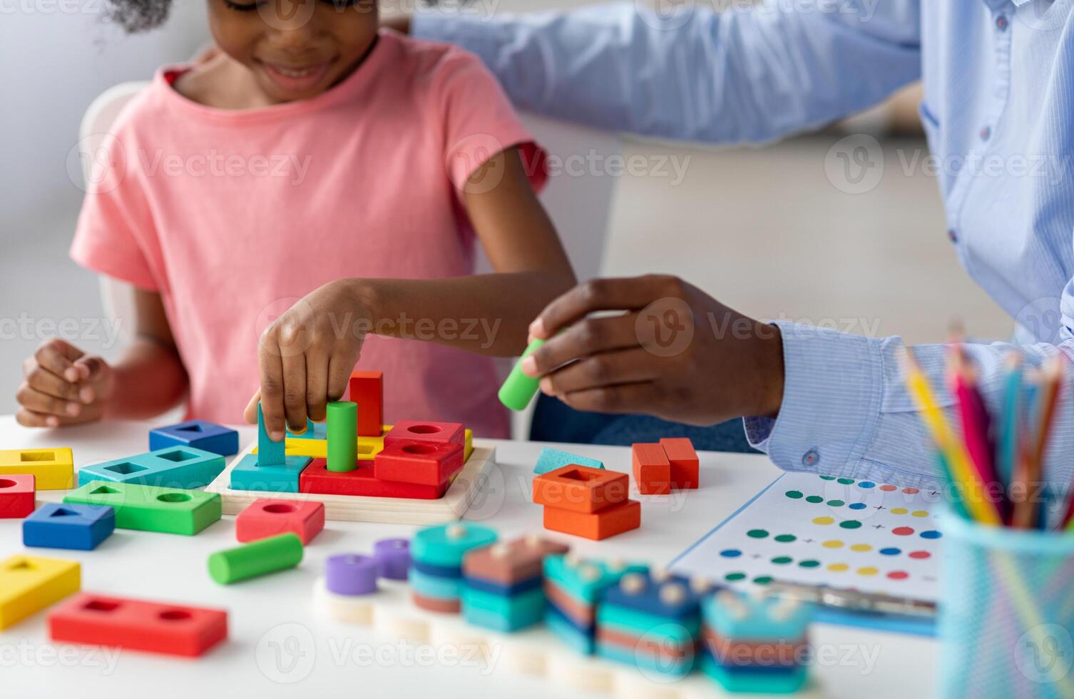 Unrecognizable little black girl with curly hair sitting at table with various wooden blocks and bricks, exercising with female teacher at daycare or attending child development specialist, cropped photo