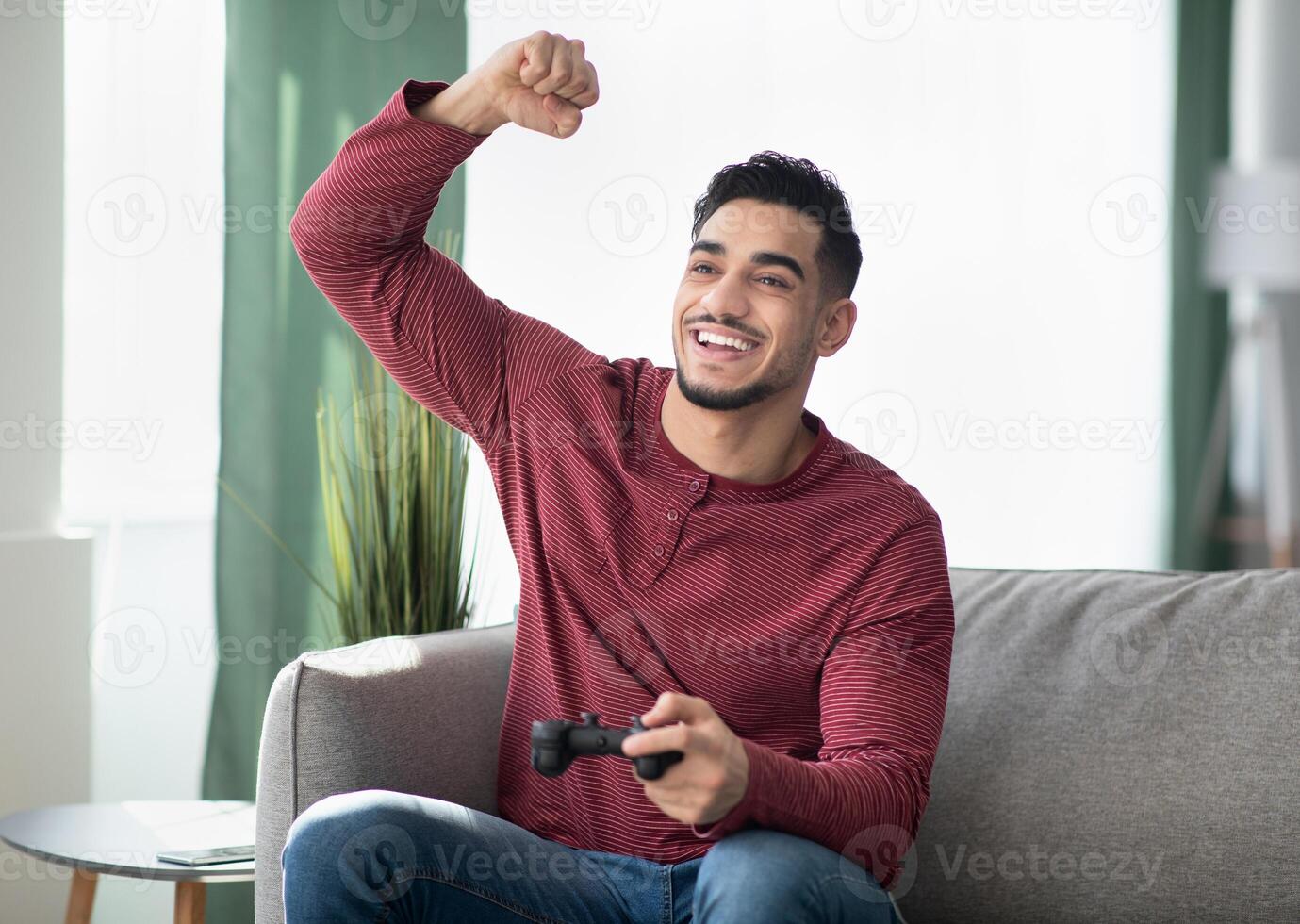 Happy millennial arab man enjoying weekend, sitting on couch in living room with joystick, gaming. Emotional middle-eastern guy playing game with joystick at home, copy space photo