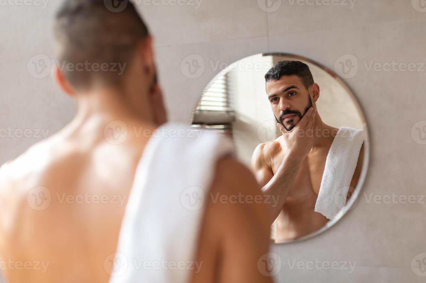 Male Facial Care. Portrait Of Handsome Arab Man Looking At His Reflection In Mirror In Bathroom And Touching Beard, Attractive Masculine Middle Eastern Guy Enjoying His Appearance, Selective Focus photo