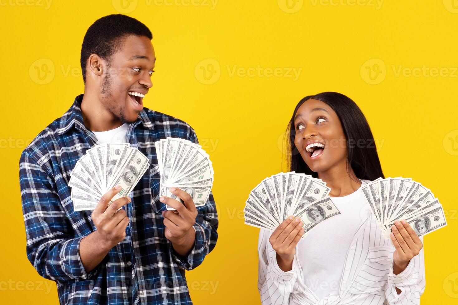 A lot of money and win. Millennial african american male and female hold many dollars in hands and expressing success at covid-19 quarantine, isolation on yellow background, studio shot, copy space photo