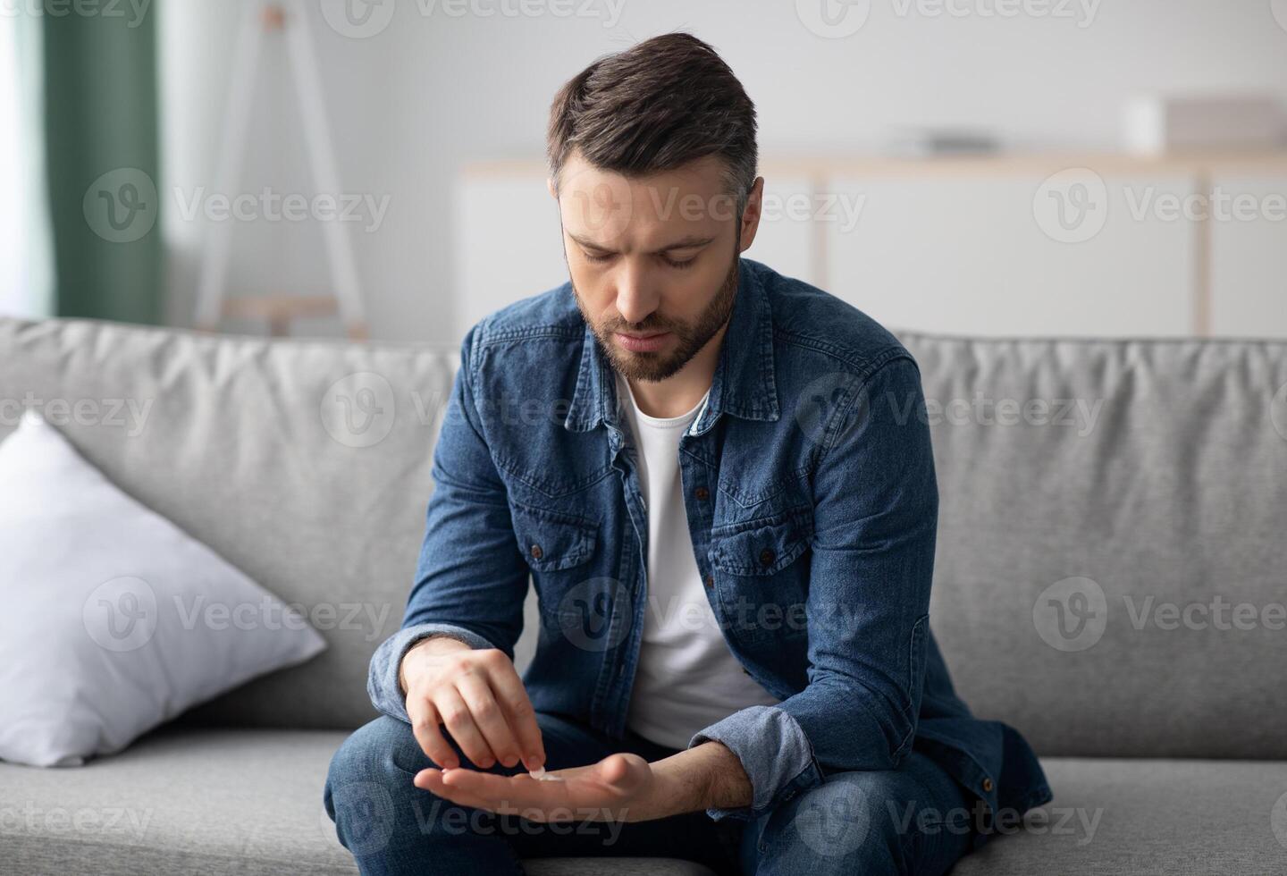 Upset middle-aged bearded mad sitting on couch in living room and looking at pills in his hands, suffering from depression or having sexual dysfunction, copy space. Medication, medicine, pills photo