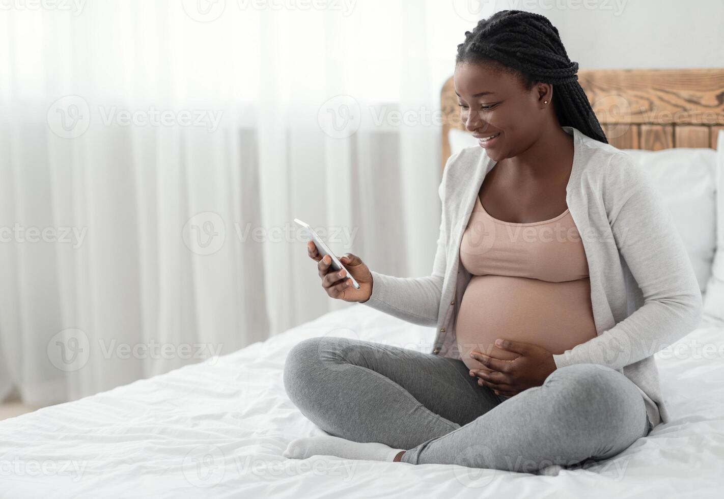 Smiling Black Pregnant Woman Sitting With Smartphone On Bed At Home, Looking At Cellphone Screen With Excitement, Happy African Lady Browsing Pregnancy App Or Received Mobile Offer, Copy Space photo