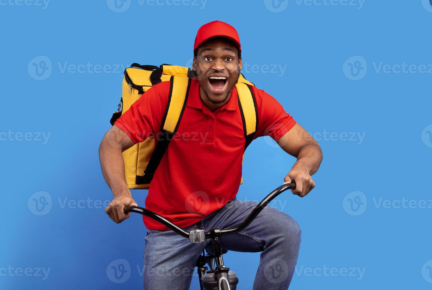 Fast Delivery Service. Overjoyed african american courier guy in red cap and uniform riding bicycle and wearing thermo backpack bag isolated on blue studio background. Shipment service during lockdown photo