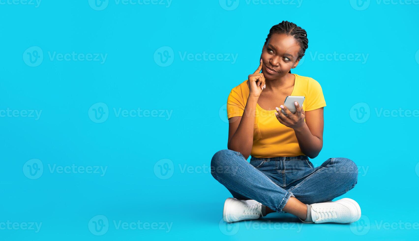 Thoughtful African American Girl Using Cellphone Sitting Over Blue Studio Background. Mobile Application Concept. Panorama With Blank Space photo
