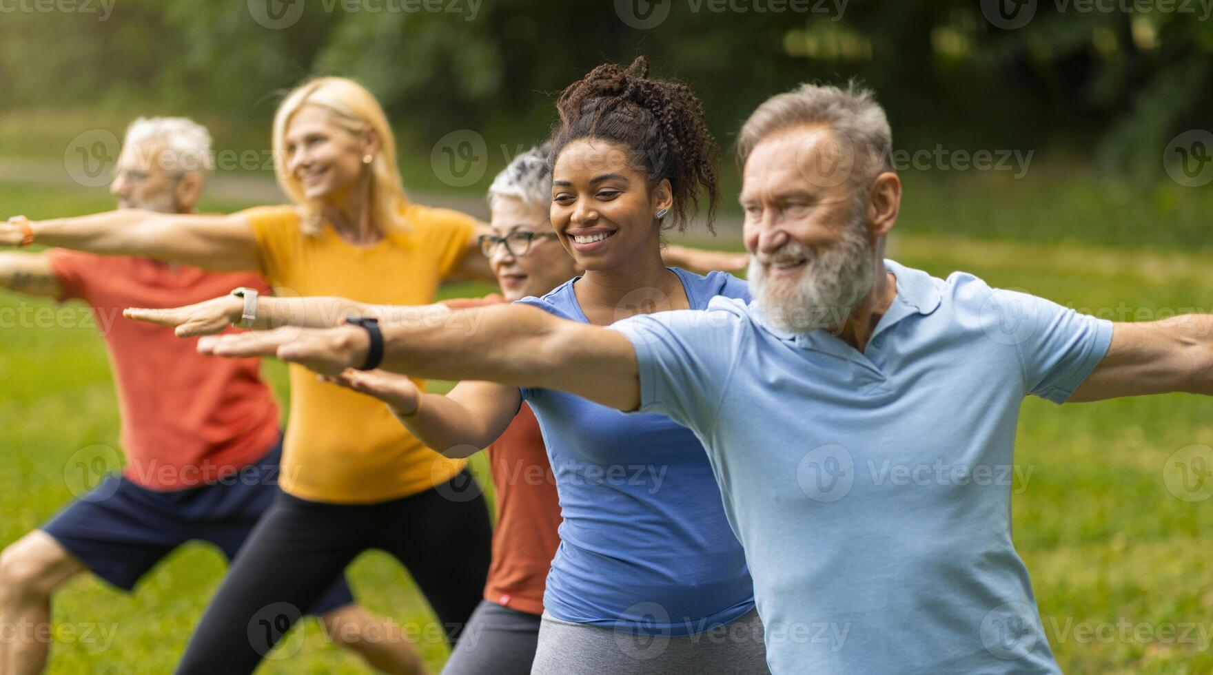 Group of diverse seniors led by young instructor practicing warrior yoga pose outdoors, happy elderly people promoting inclusivity and wellness, exercising in lush park setting, closeup shot photo
