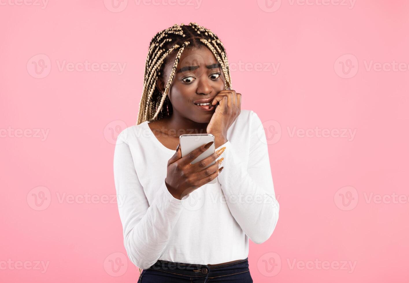 Portrait of anxious scared black young woman looking at mobile phone seeing bad news, photos or message. Worried lady biting nails isolated on pink studio wall background. Human reaction, expression