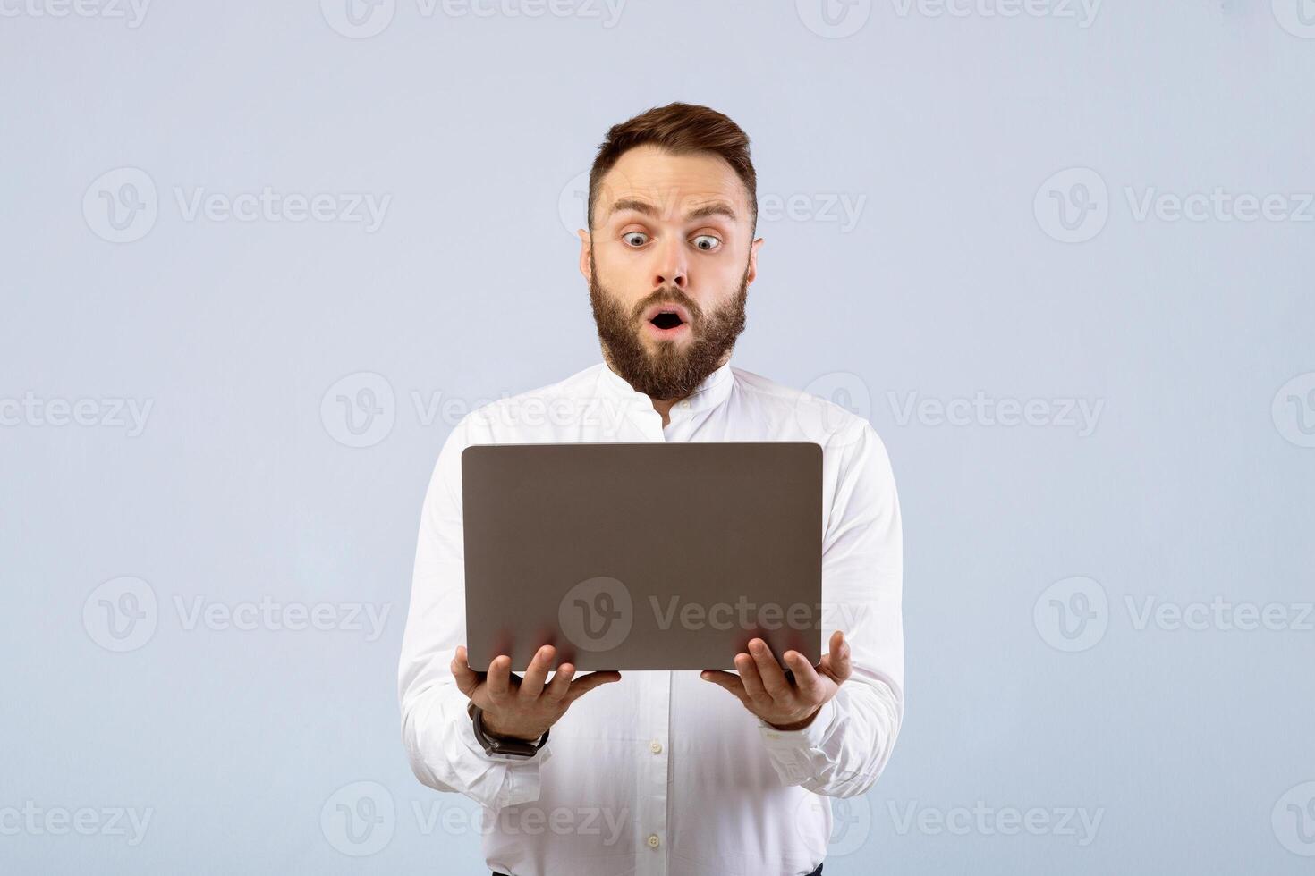 Shocked young businessman in formal wear looking at laptop screen in disbelief on grey studio background. Stunned office worker learning unbelievable news online, learning about huge sale photo