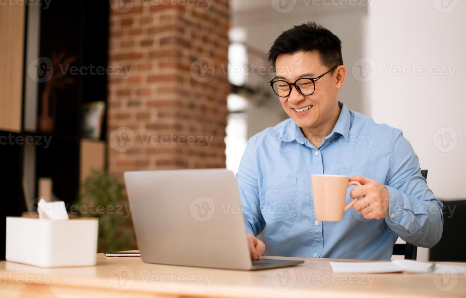 Asian middle aged man drinking coffee at home office, sitting at workplace with computer, free space. Business, coffee break from work, pause and vitality for freelance photo