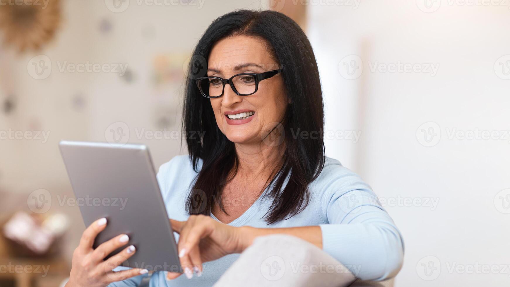 Portrait Of Smiling Mature Woman With Long Dark Hair Wearing Eyeglasses, Holding And Using Digital Tablet, Watching , Reading E-Book Or Making Online Call, Surfing Internet Sitting On Sofa photo