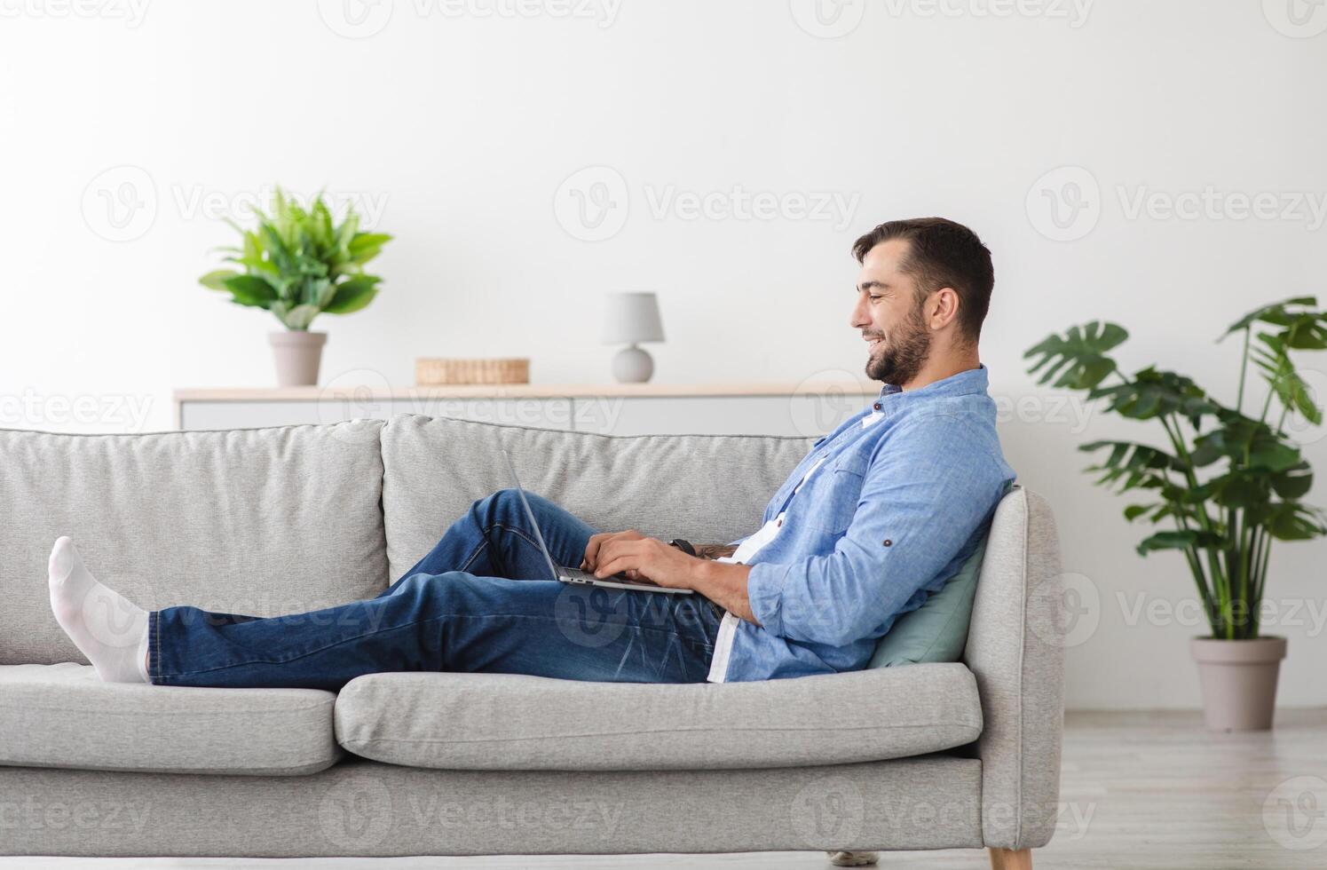 Happy adult european man with beard sitting on sofa and typing on laptop in living room interior, profile, copy space. Guy plays in online game at free time, chat on social networks and work remotely photo