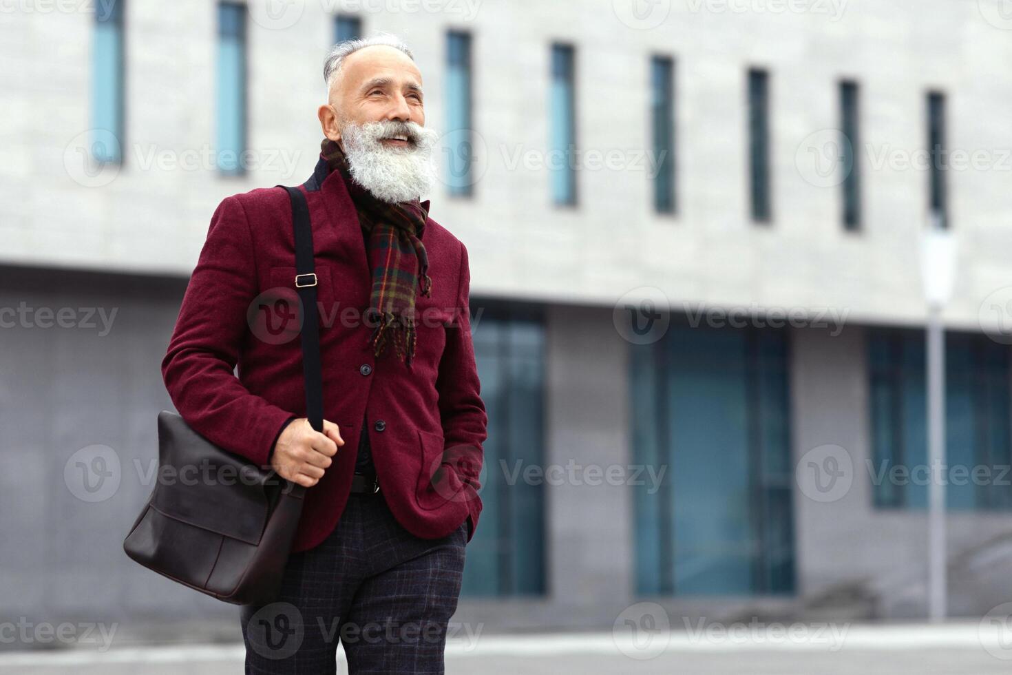 Happy senior grey-haired man in stylish burgundy jacket walking by street alone, going home from job, smiling and looking at copy space. Working during retirement concept, lifestyle of elderly photo