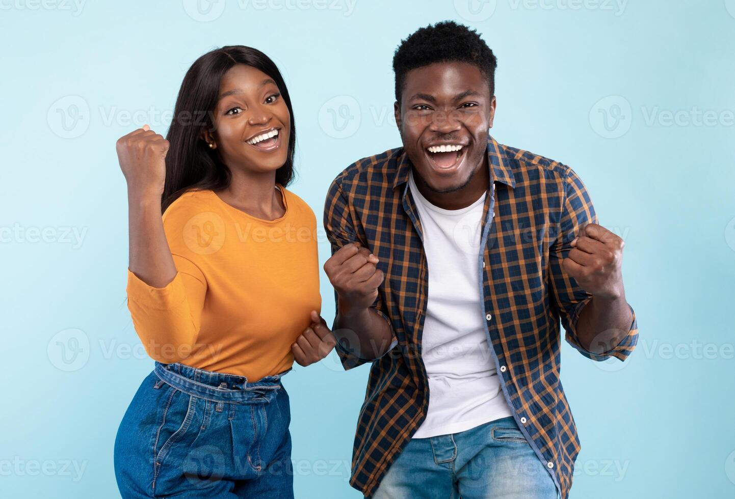 Wow, Yes. Portrait of overjoyed young black man and woman cheering and shaking clenched fists, looking at camera. Happy couple celebrating win posing standing isolated over blue studio background wall photo