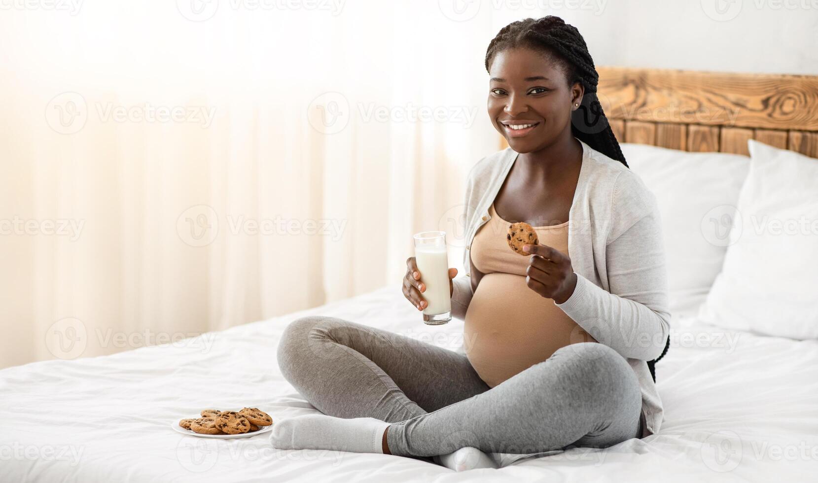 Snack Time. Happy Black Pregnant Woman Eating Cookies And Drinking Milk At Home, African Expectant Lady Having A Bite While Relaxing On Bed In White Bedroom, Looking At Camera And Smiling, Copy Space photo