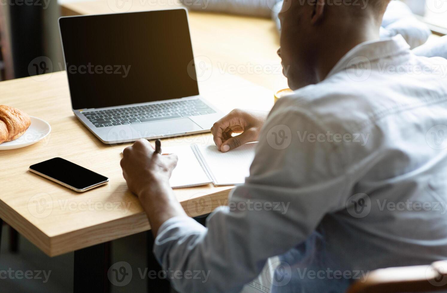 Unrecognizable black guy using laptop with blank screen, studying online, taking notes in copybook at cafe, mockup for design. Millennial man working remotely at coffee shop, space for website photo