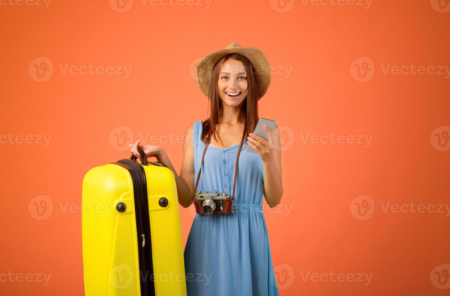 Happy young woman holding suitcase and using mobile phone over yellow studio background, found online great deals for travelling. Active resting, vacation, tourism, travelling concept photo