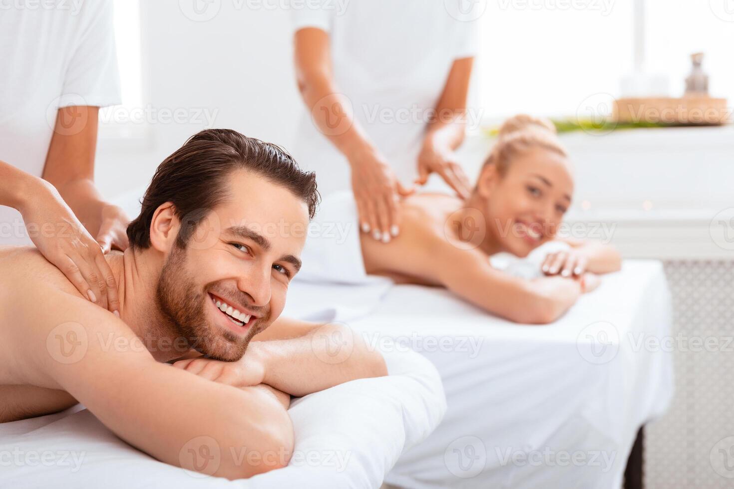 Happy Couple Receiving Back Massage At Spa Salon. Spouses Enjoying Beauty Treatment Together Smiling To Camera Lying On Beds Indoors. Honeymoon Vacation Relaxation, Romantic Wellness Procedure photo