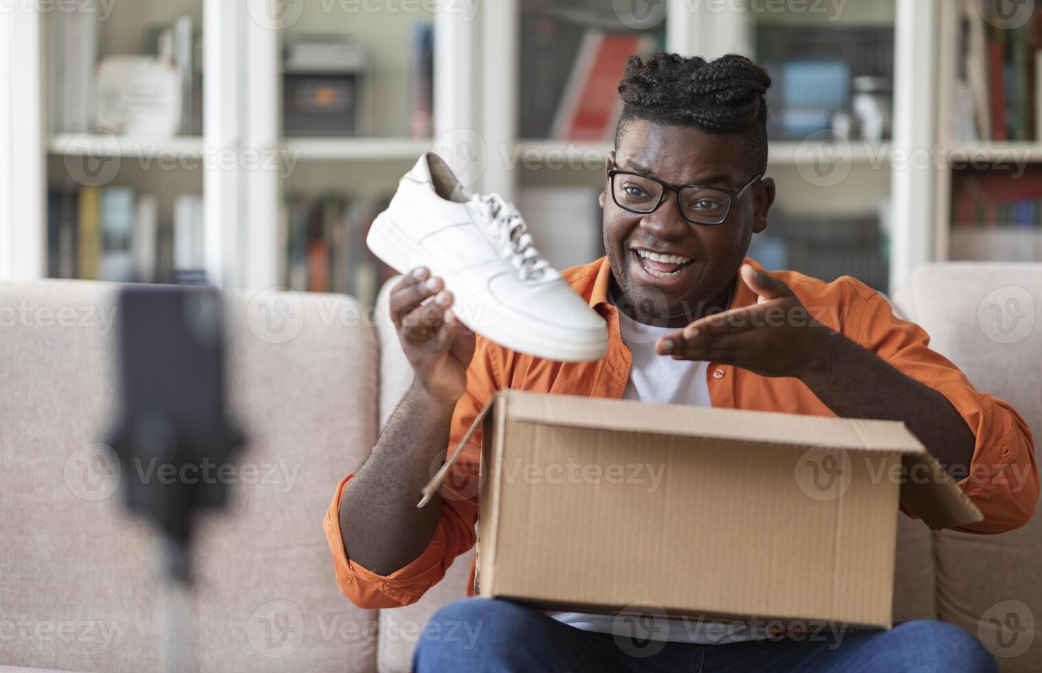 Social media influencer stylish cheerful chubby black guy reviewing product. Excited african american millennial man vlogging about unpacking white sneakers and filming himself at home on cell phone photo