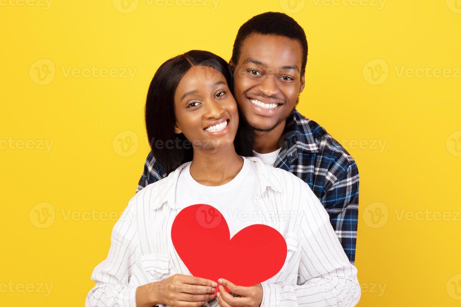 Romantic relationships and Valentines day during self-isolation. Cheerful young african american couple in love hug and hold red heart and look at camera, isolated on yellow background, studio shot photo