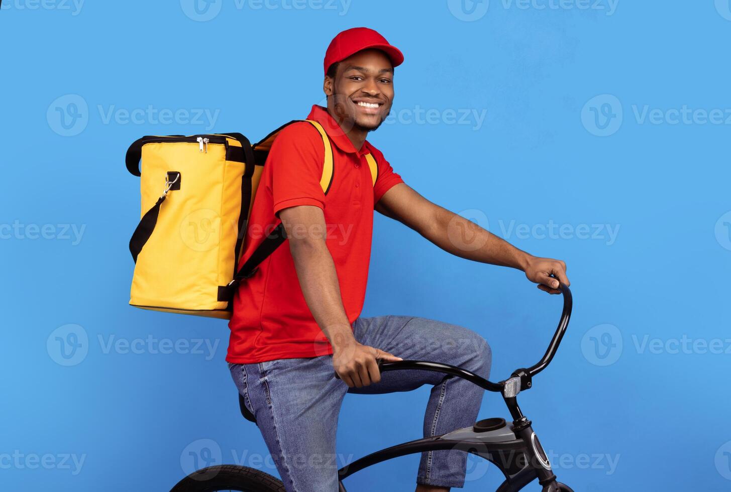 Delivering Orders On Time. Portrait of cheerful black male courier in red cap and uniform riding bicycle and wearing yellow thermal backpack bag with takeaway food posing at blue studio background photo