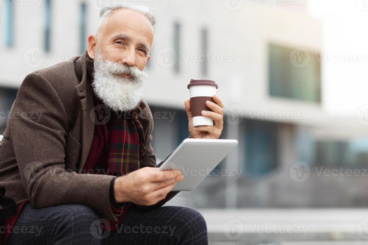 Handsome aged grey-haierd man holding paper cup with coffee and using modern digital tablet, sitting on street during break, copy space. Elderly man drinking morning coffee and using pad photo