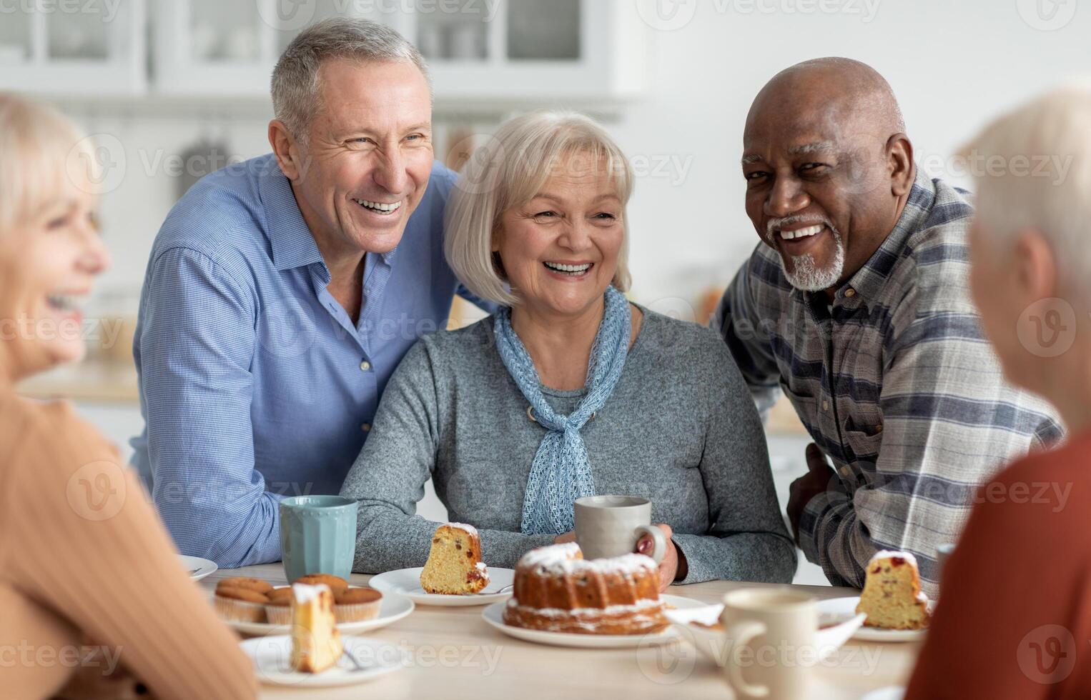 Cheerful senior people of different nationalities sitting at kitchen, drinking tea and eating cake together, having conversation and laughing, chilling together at nursing home photo