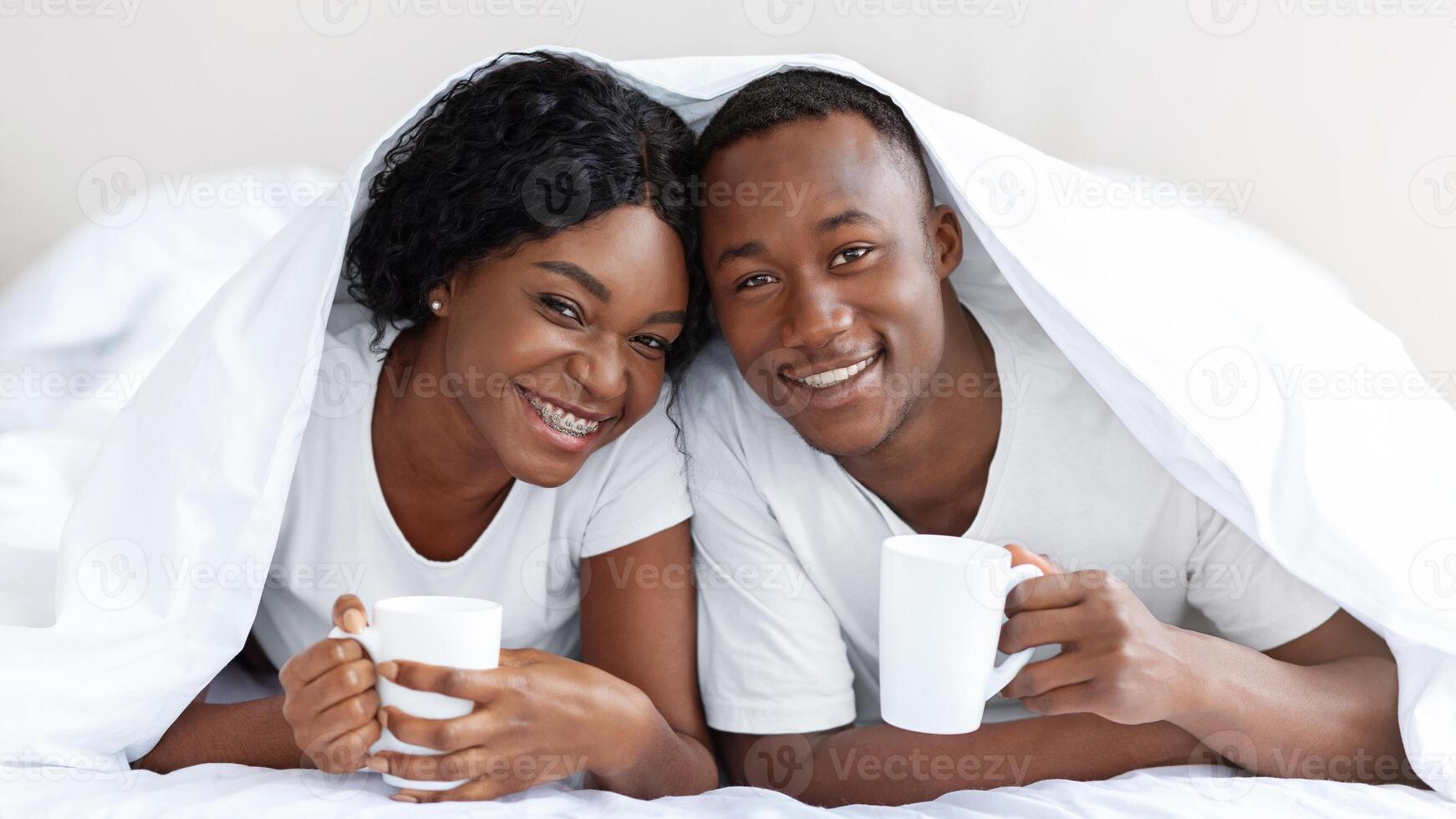 Happy black couple laying under blanket, drinking hot tea, warming up during cold weather. Cheerful african american lovers with mugs staying in bed during cold winter day, bonding photo