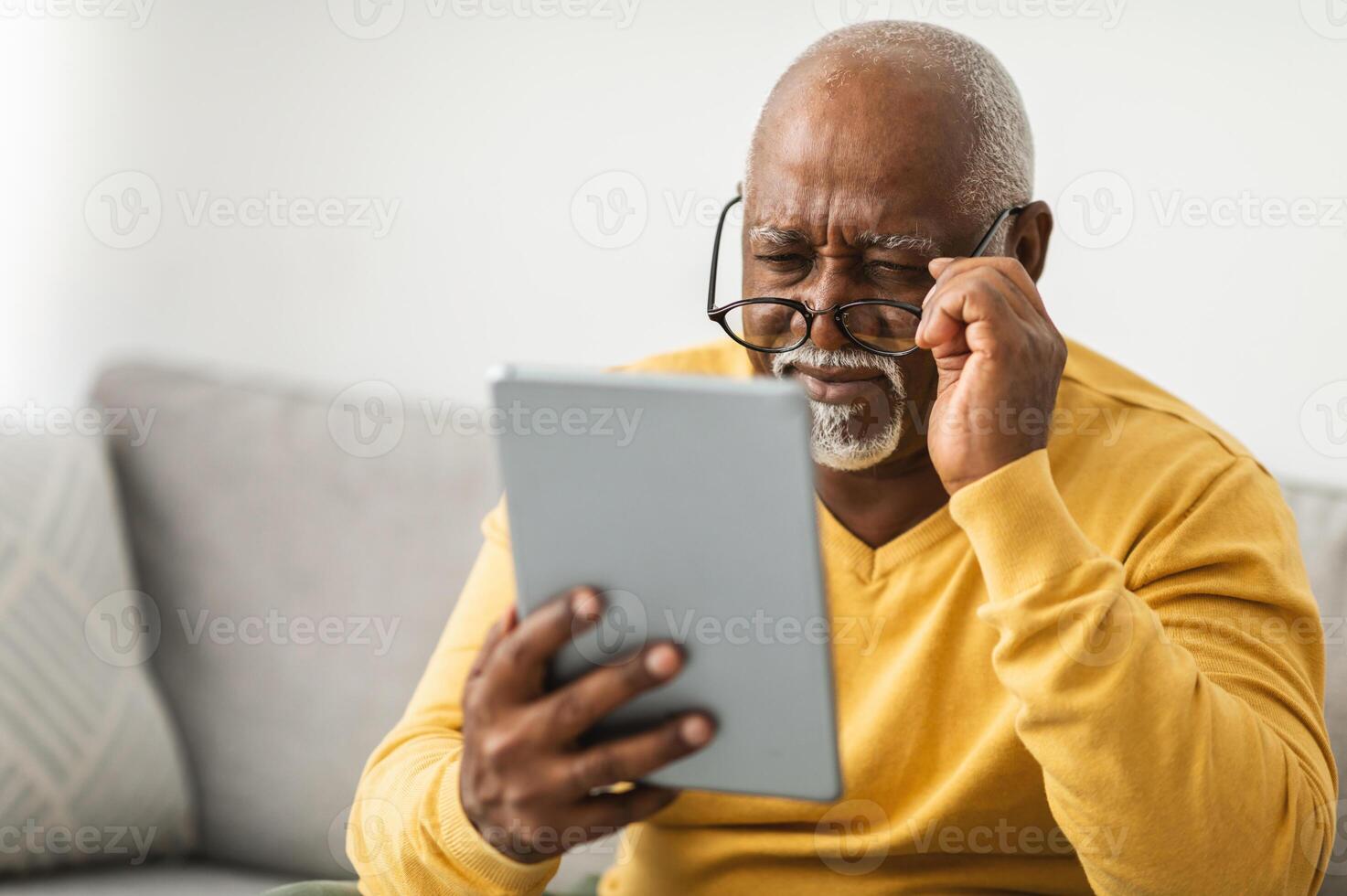 Senior Black Man With Poor Eyesight Using Digital Tablet Wearing Glasses And Squinting Eyes Reading News Online Sitting On Couch At Home. Eyes Health Issues Concept. Selective Focus photo