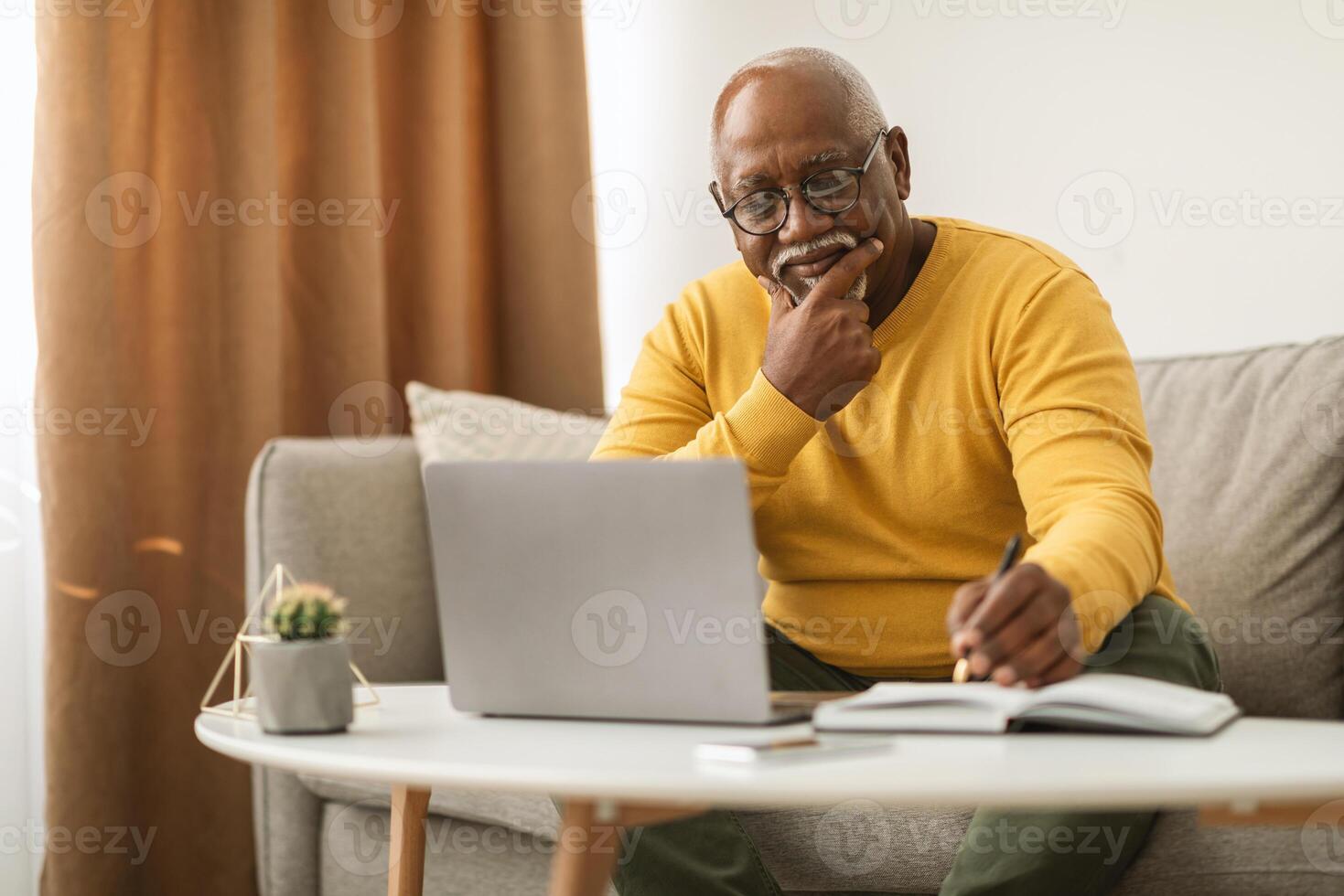 Mature African American Business Man Using Laptop And Writing Taking Notes Sitting On Couch Indoor. Senior Male Freelancer Working Online On Computer At Home. E-Learning In Older Age photo