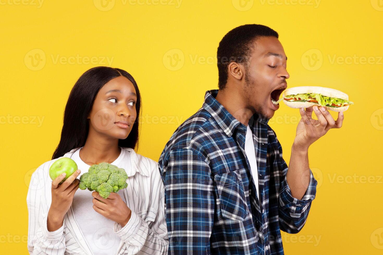 Healthy nutrition and fast food, diet and relationship. Funny millennial african american guy eats sandwich, wife with vegetables and fruits sadly looks at husband isolated on yellow background photo