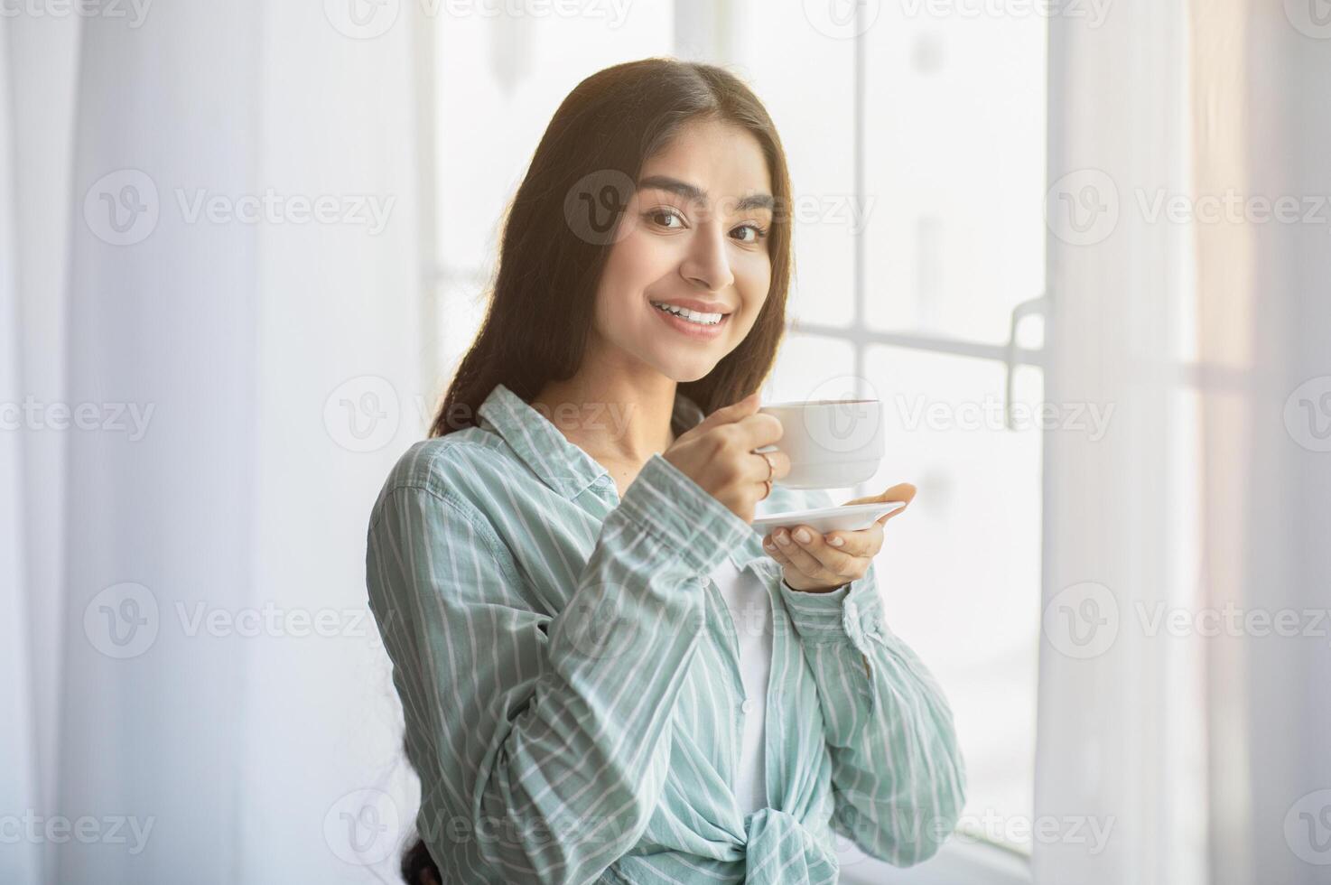 Lovely young Indian woman having coffee break in front of window at home. Charming millennial lady relaxing, enjoying lazy weekend morning with hot drink, smiling at camera photo