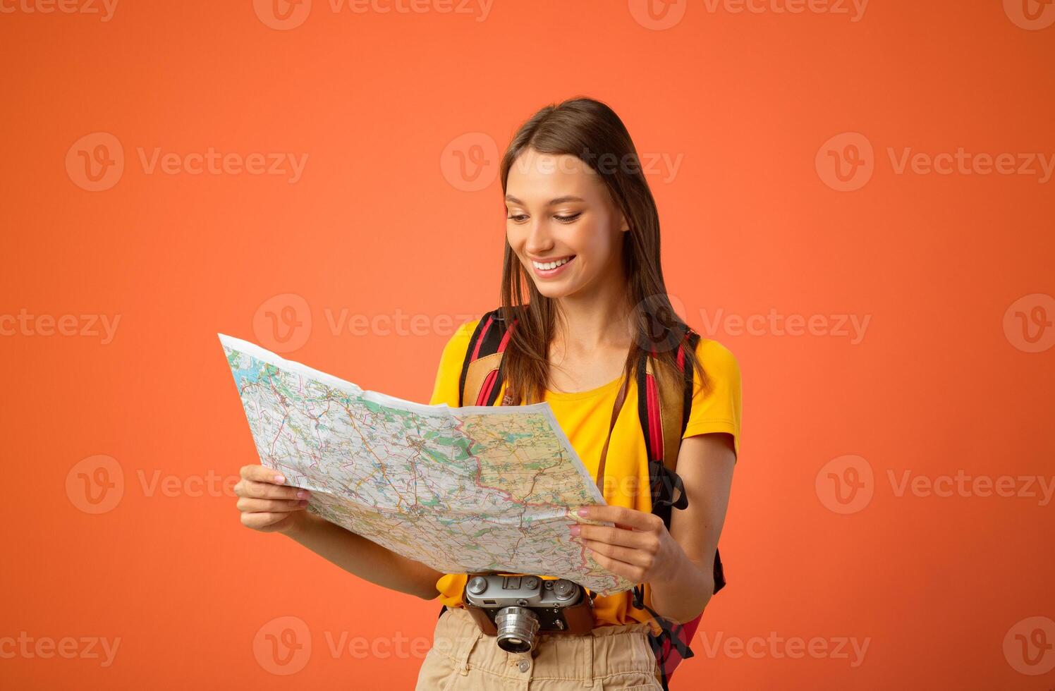 Happy young woman travelling alone, holding city map, having camera and backpack, yellow studio background, copy space. Joyful female traveler looking for new place to go, travelling concept photo
