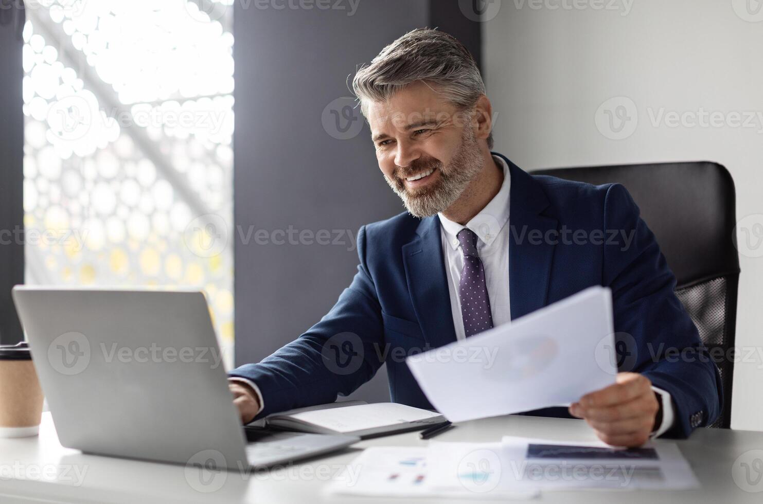 Successful middle aged entrepreneur working with laptop and papers at workplace in modern office, smiling mature businessman in suit sitting at desk and managing financial reports, copy space photo