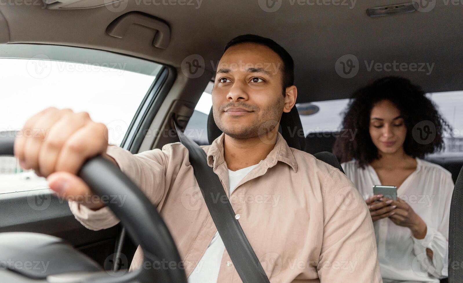 Confident Indian Taxi Driver Navigating Car With Passenger Woman, Driving Skillfully In City Traffic, Lady In Backseat Using Cellphone During Automobile Ride. Selective Focus. Cab Transportation photo