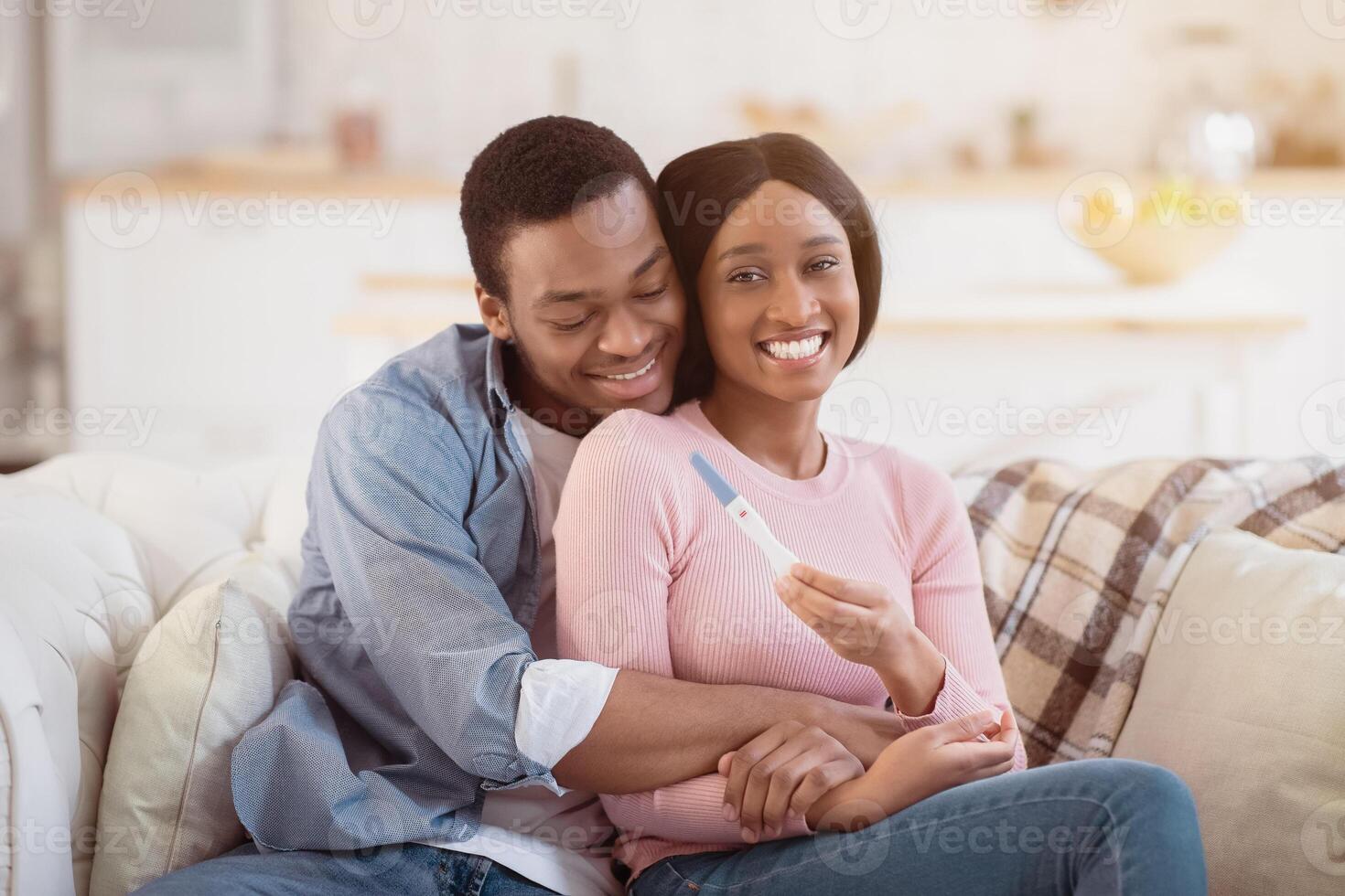 Young African American family learning about their future child from pregnancy test at home, embracing on sofa. Loving husband and his beautiful expectant wife cuddling and feeling happy photo