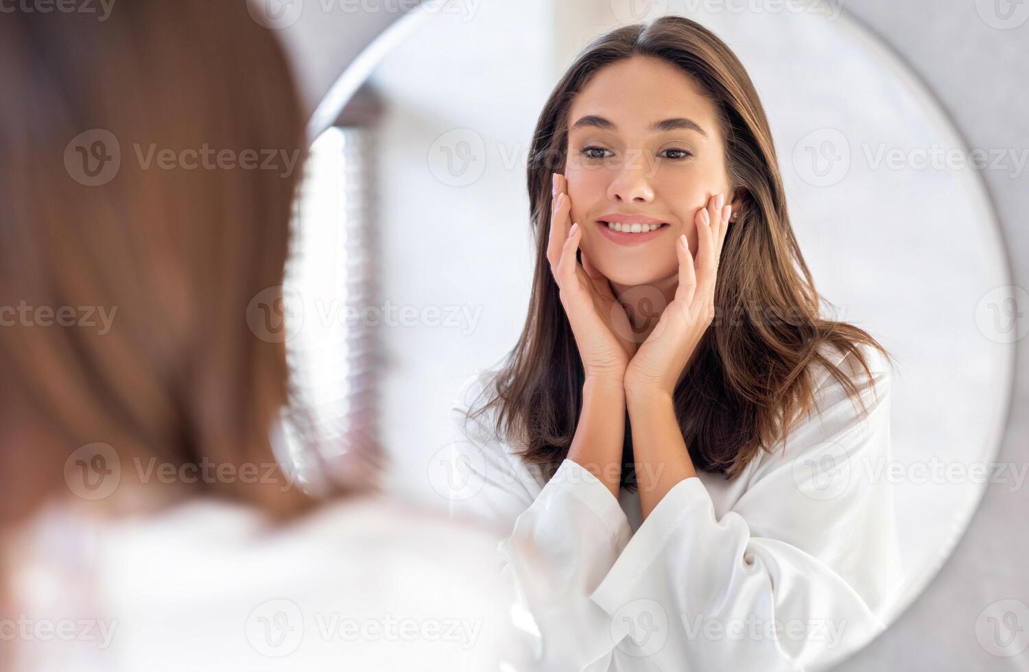 Skincare Concept. Pretty Young Woman Touching Her Face While Looking At Mirror In Bathroom, Attractive Female Smiling To Reflection, Enjoying Skin Condition After Beauty Treatments, Selective Focus photo