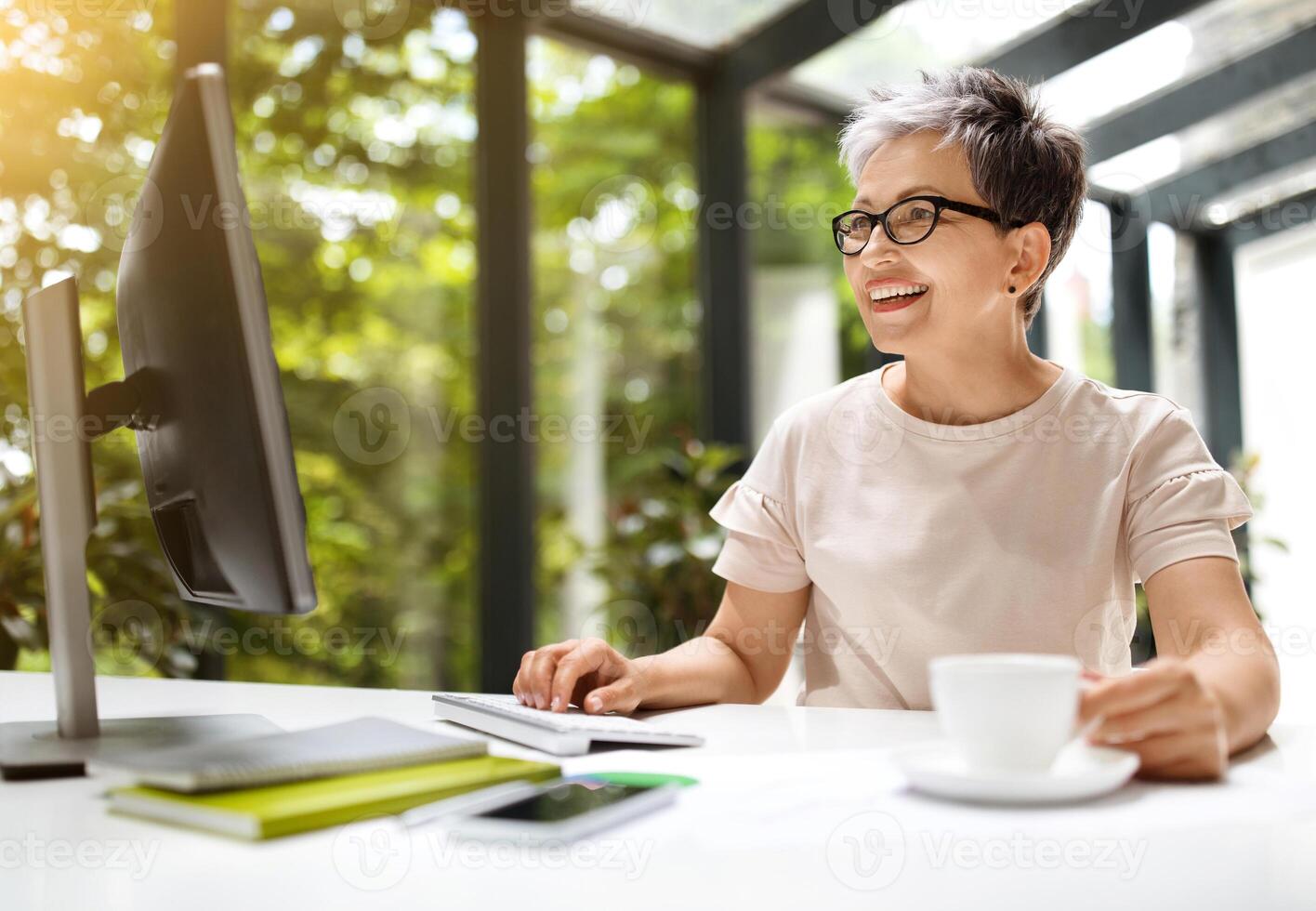 Happy beautiful stylish mature woman wearing eyeglasses employee sitting at workdesk, using pc computer, sending emails, drinking coffee, working at coworking space, copy space, sun flare photo