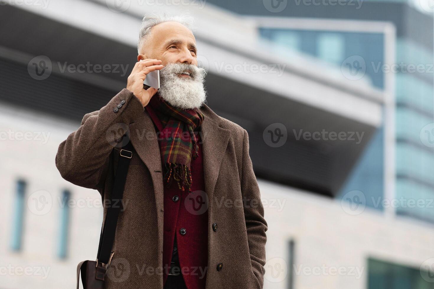 Wealthy senior grey-haired man with long beard having conversation on mobile phone while walking by street, standing by business center, going to business meeting, looking at empty space photo