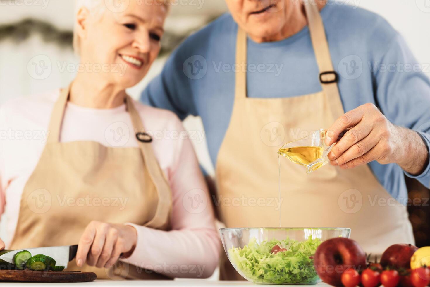 Unrecognizable Senior Couple Cooking Adding Olive Oil To Salad Preparing Dinner In Modern Kitchen At Home. Elderly Spouses Enjoying Food Preparation. Cropped, Selective Focus, Shallow Depth photo