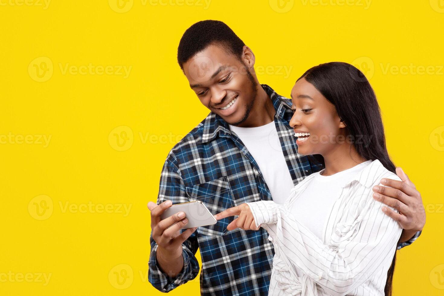 Amazing message about promotions, discounts, sales and online app. Smiling millennial african american students guy and lady look at phone, female controls finger, isolated on yellow background photo
