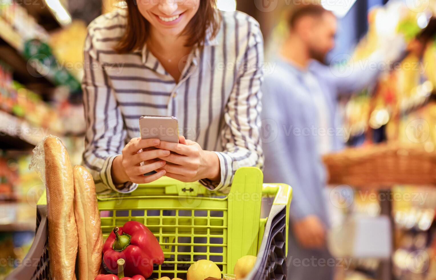 Couple In The Supermarket. Cropped closeup image of unrecognizable smiling woman leaning on shopping cart, using mobile phone, her boyfriend is choosing food in the blurred background photo