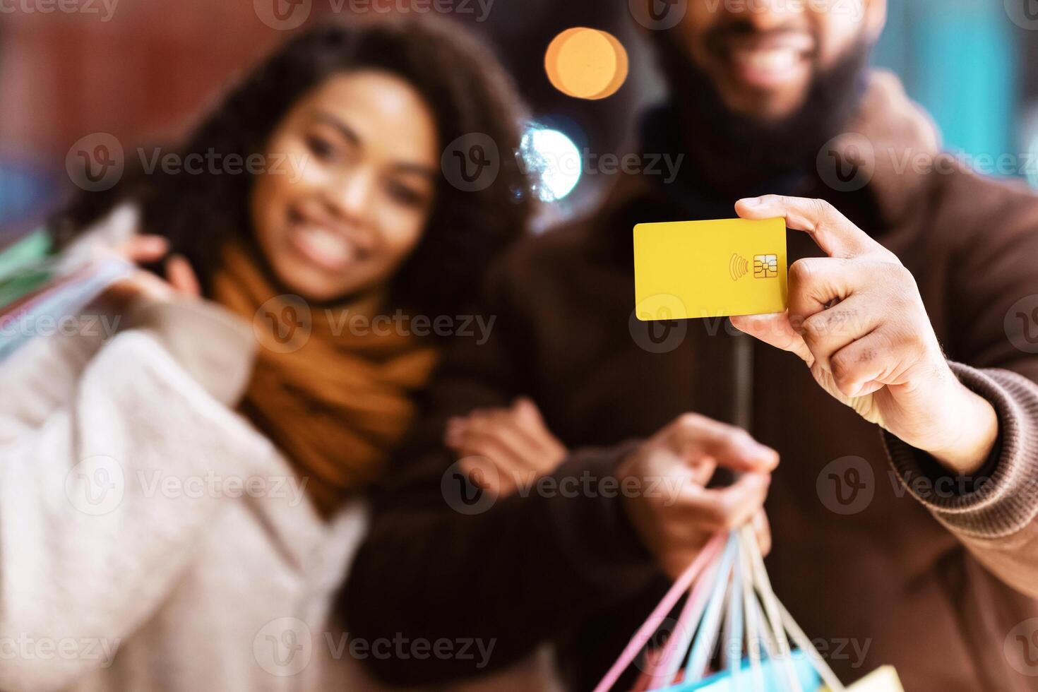 Easy Shopping. Portrait of smiling black couple showing debit credit card and holding shopper bags, walking outside in the city near mall in the evening, selective focus, blurred background photo