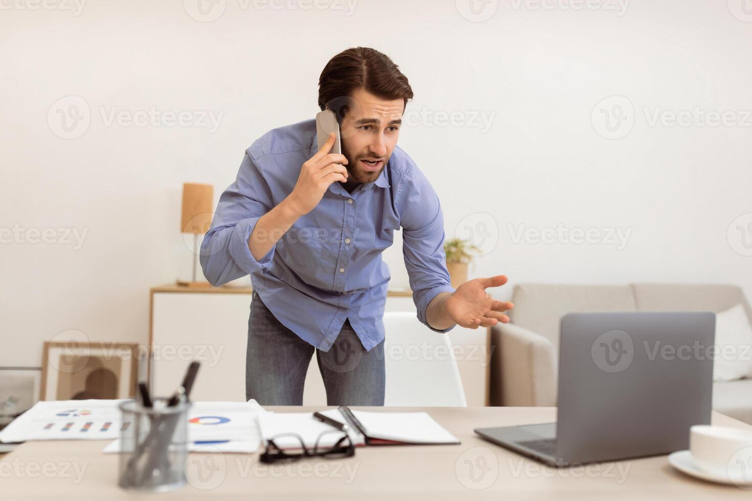 Displeased Businessman Talking On Cellphone Having Computer Problem Standing In Modern Office. Discontented Customer, Communication Issue, Problems At Work Concept. Selective Focus photo