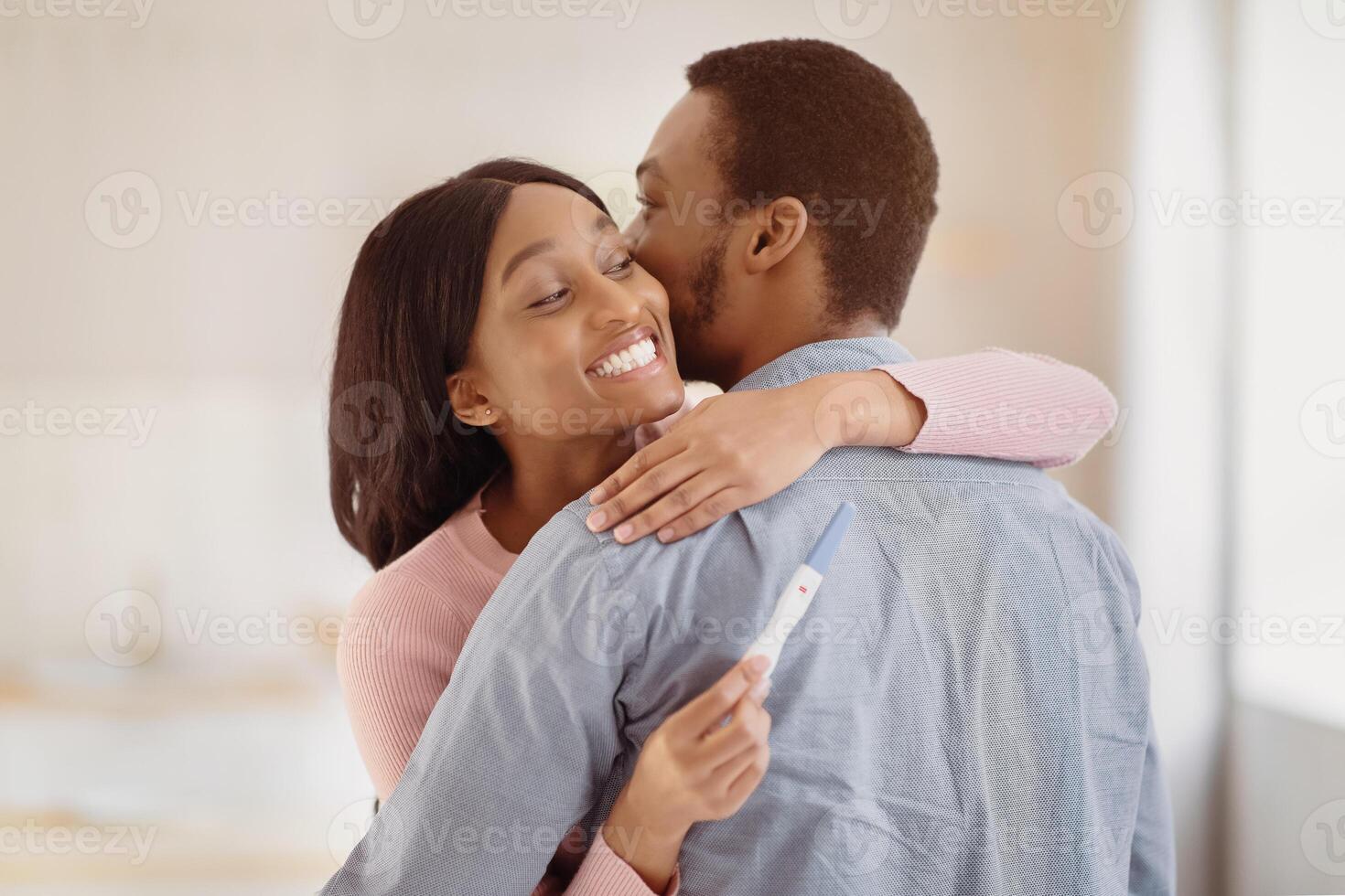 African American man embracing his happy young wife with positive pregnancy test indoors. Millennial black couple expecting baby. Maternity and childbearing concept photo